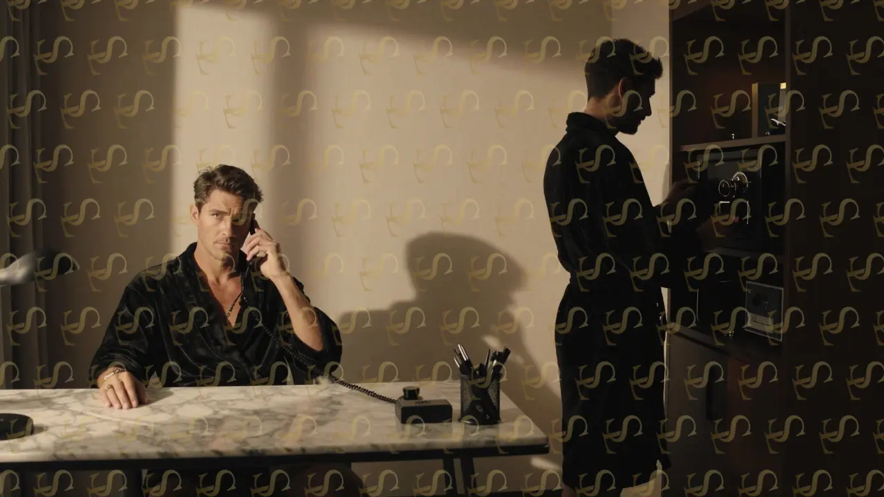 Two figures in robes near minibar and desk, sharing a quiet corporate moment in dim light.