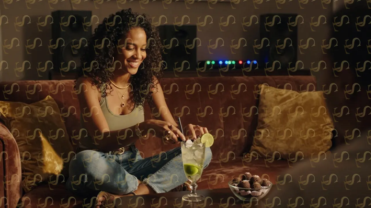 A Brazilian companion teaching a client to make a caipirinha in a luxurious Dubai living room, brigadeiros on the table.